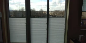 Interior view of large glass doors with white horizontal blinds, outside showing bare trees and a cloudy blue sky. When closed, blinds obscure most of the view to the outside.